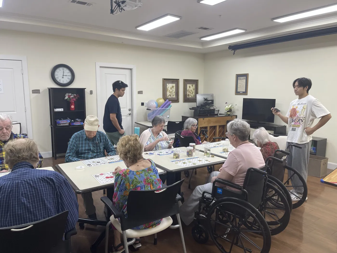Two student volunteers stand among residents playing bingo at tables in an activity room